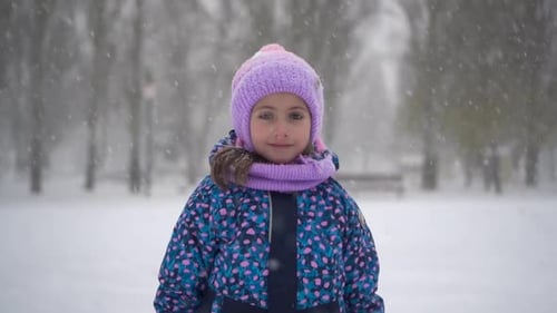 A Little Girl Stands Under Snowfall in the Park A Girl in a Jacket and Hat Looks at the Camera Slow