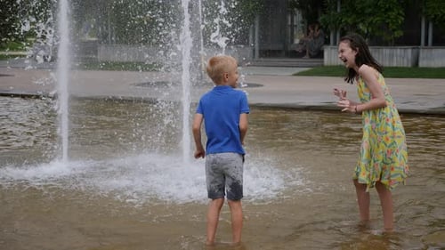 Two Cheerful Children Play in the Fountain in the Summer in the City Park
