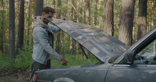 Young Man Driver Fixing Car Under Hood Repairing Automobile Trouble During Road Trip in Forest