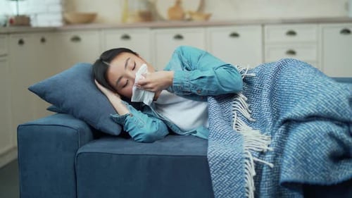 Une jeune femme asiatique au rhume et au nez qui coule est allongée sur le canapé recouverte d'une couverture
