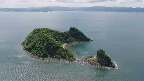 Aerial orbit reveals secluded tropical island with dense forest canopy, cliffs and hidden cove. Pris