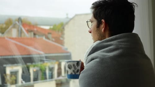 Man Wrapped in Blanket Drinking Coffee by Window