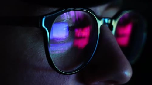 Man on his computer at night ultra violet blue light hitting glasses - closeup handheld shot