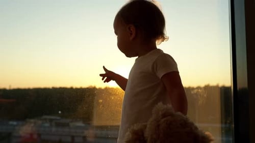 Child Holding Teddy Bear Standing By Window At Sunset