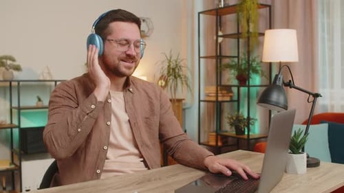 Happy Caucasian Man Using Laptop While Listening to Music Via Headphones Sitting at Home Office Desk