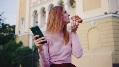 Woman Eating Croissant While Using Mobile Phone Outdoors