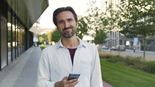A Caucasian Man Looks at a Smartphone and Then Laughs at the Camera in a Street in an Urban Area