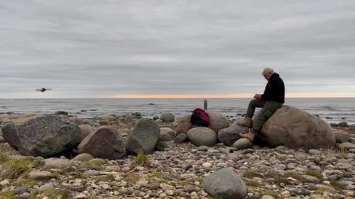 Man operating drone while sitting on rocky shore near ocean