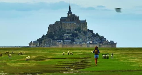 A Female with a Backpack Visits the Mont Saint Michel Castle While Walking Through Green Meadows
