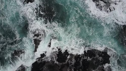 Aerial: Turbulent ocean waves crashing over dark basalt rock ledges. Camera looking directly down wi