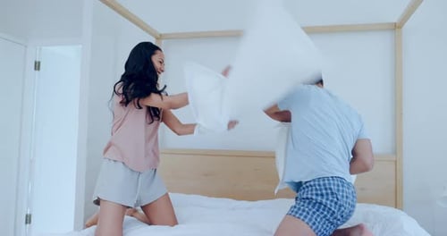 Happy couple, bed and playing in pillow fight, bonding or fun holiday weekend together at home