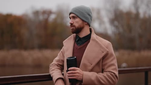 Pensive Man in Autumn Coat and Beanie Drinking Coffee by the River