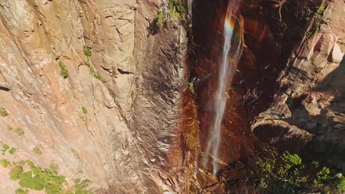 Rainbow appearing in the stream falling from huge cliff. Drone footage raising along the steep rocks