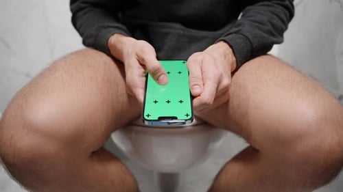 A Man with His Pants Down Is Sitting on the Toilet and Scrolling Through Photos on a Smartphone.