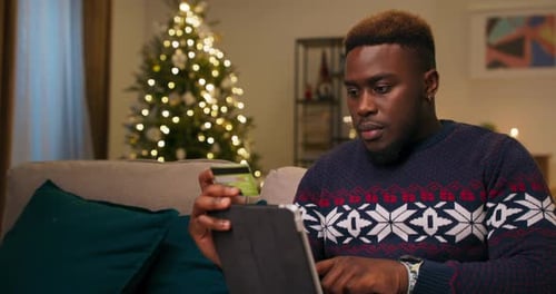 A Young Man of African Appearance Sits on Sofa in the Living Room Holding Tablet and Bank Card and