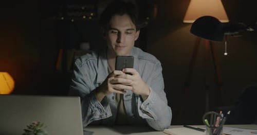 Portrait of Joyful Young Man Using Smartphone Then Looking at Camera and Smiling in Dark Office at