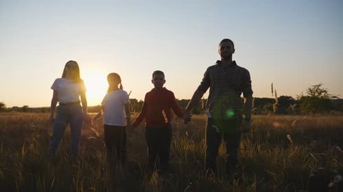 Happy Family Looks Into Camera Standing on Grass Field and Holding Hands of Each Other at Sunset
