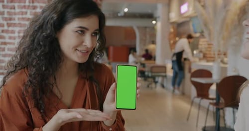 Young Woman Showing Green Screen Chroma Key Smartphone to Female Friend in Cafe Talking and Laughing