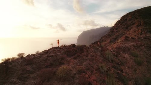 Person in red stands on a rocky hill, arms spread wide, taking in the view of the ocean and cliffs d