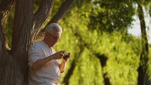 Senior Man Uses Smartphone Under Shady Tree