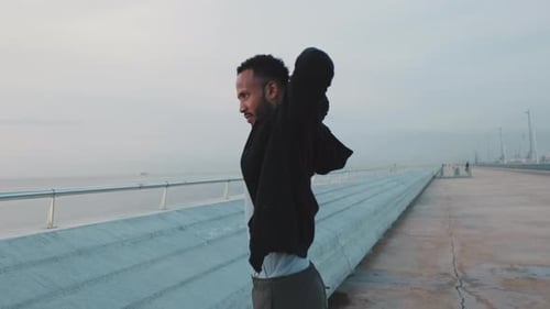 Young athlete doing warm-up, stretching his arms before training while standing on the embankment
