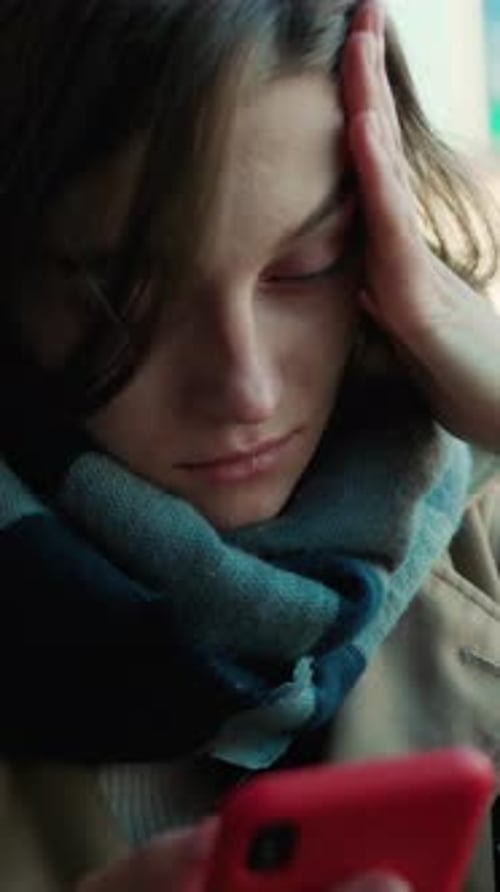 A Contemplative Woman Wrapped in a Scarf is Using Her Phone While Sitting Thoughtfully