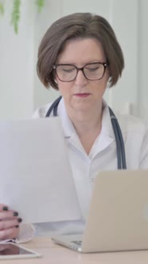 Vieille femme médecin travaillant sur des documents et un ordinateur portable en clinique, Vidéo verticale