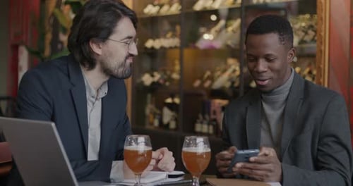 Young Men Colleagues Talking Looking at Smartphone Screen and Laughing Relaxing in Cafe Together