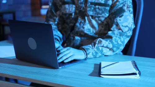 Military man working on laptop at wooden desk in office at night, closeup