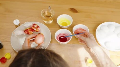 Overhead view of woman painting a pink Easter egg, with wrapped eggs and bowls of dye spread out on