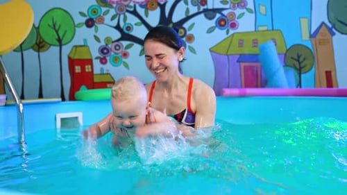 Smiling Caucasian dark-haired woman holding a little blond baby in the swimming pool.