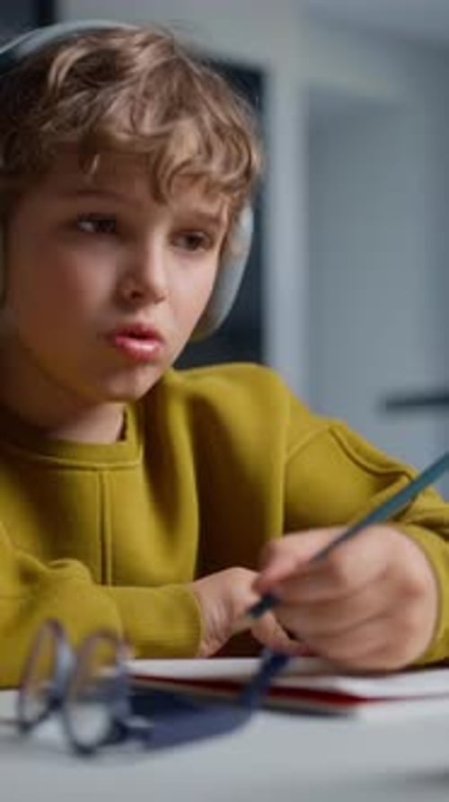 Focused Young Boy with Curly Hair Wearing Headphones and a Mustard Yellow Sweater Engaged in Writing