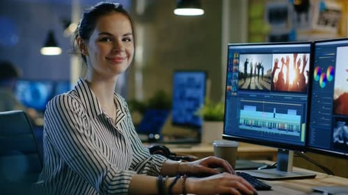 Female Video Editor Works with Footage and Sound on Her Personal Computer, She Turns and Warmly Smi
