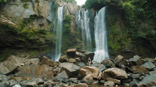 Woman walking on rocks before tall tropical waterfall surrounded by rocky cliffs