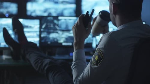 Security officer is relaxing at a computer desk while drinking coffee in a dark monitoring room fil