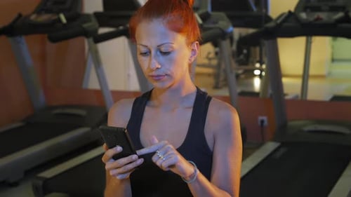 Red hair pretty girl looks photos at mobile phone after hard training in GYM