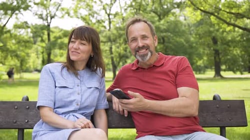 A Middleaged Caucasian Couple Listens to Music on a Smartphone As They Sit on a Bench in a Park