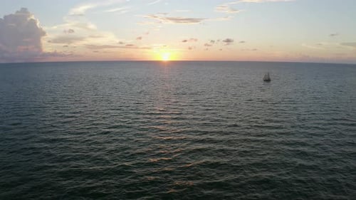 Sailboat drifting across calm ocean waters under glowing sunset horizon — aerial view