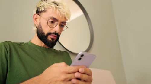 Close-up, young blond man with a beard sits on a sofa in a cozy living room using a mobile phone