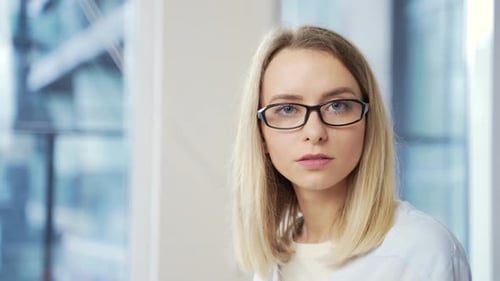Close-up portrait of a young adult blonde business woman with glasses looking at the camera indoors