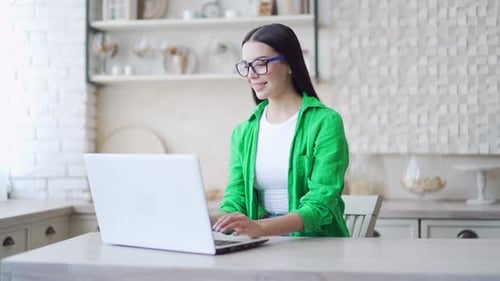 Smiling brunette with eyeglasses using laptop in the light room. Attractive woman in green shirt