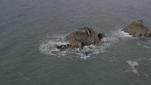Aerial of SUP athlete paddling in chaotic water near rocky ocean islet