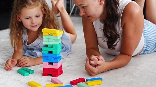 Little Girl with Her Mother Playing Jenga Board Game at Home Taking Colorful Blocks From Wooden