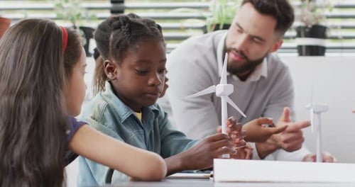 Diverse male teacher and happy schoolchildren studying wind turbines in school classroom