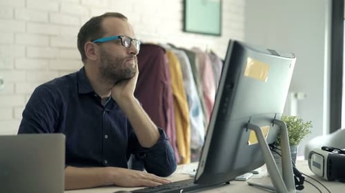 Junger gelangweilter Mann checkt Laptop und Smartphone und sitzt zu Hause am Tisch der 30er