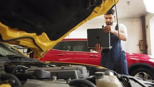 Close Up of Young Bearded Engineer Mechanic Under Car Hood Checking Engine with
