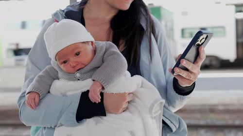 Mother holding a newborn baby while checking her phone at a train station. the care and attention of