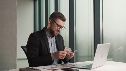 Um jovem feliz com roupas de negócios conta contas de dinheiro enquanto está sentado em um laptop