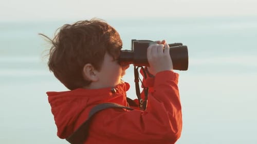 Happy boy looks through big binoculars into distance of sea against sunset