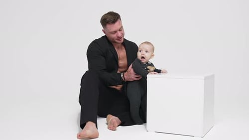 Strong muscular man in black jacket on bare chest holds a baby. Father and son studio footage.